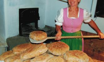 A baker in traditional dress stands in front of a wood oven and holds bread with a shovel. In front of her lies a stack of freshly baked bread.