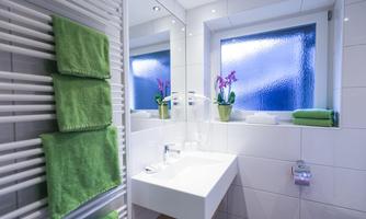 A modern bathroom with white tiles and a large mirror. Green towels and a plant provide a fresh accent.