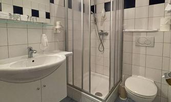 A small bathroom with a shower, a sink, and a toilet. The walls are light-tiled, and there is a shelf for toiletries.