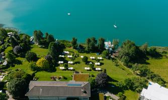 A green meadow by the water with sun umbrellas and trees. In the background, boats can be seen on the blue lake.
