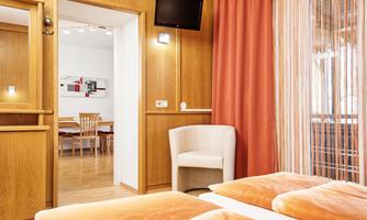 A cozy room with wood paneling and a large window. In the background, a dining area can be seen.