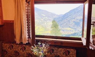 A cozy room with a wooden table and flowers in a vase. Through the open window, one looks out at a picturesque mountain landscape.
