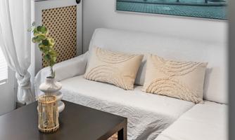 A cozy living room setup with a white sofa, decorative cushions, and a small table. In the background, there is a large picture and plants provide fresh accents.