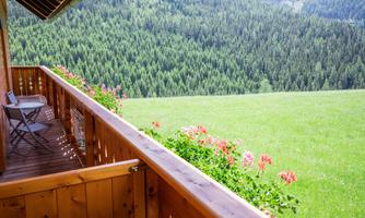 A balcony with a wooden railing, adorned with flowers. In the background, green meadows and wooded mountains stretch out.