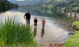 A family stands in the shallow water of a lake. In the background, green hills and trees can be seen.