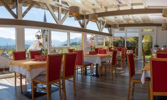 A bright restaurant with large windows and a beautiful view of the mountains. The tables are decorated with white tablecloths and red chairs.