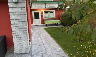 A red painted house with a green terrace. The path is paved and there are lawn areas and plants in the garden.