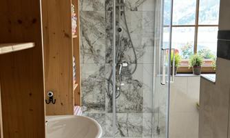 A modern bathroom with a glass shower and stylish marble. In the background, a sink and windows with plants can be seen.