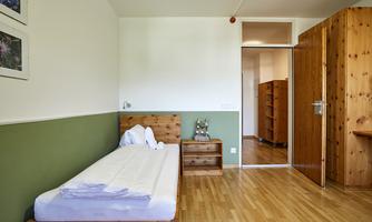 A simple room with a single bed and wooden furniture. The walls are green and there is a wardrobe as well as a table.