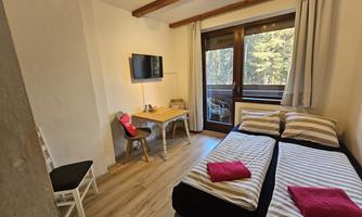 A cozy bedroom with two beds and colorful pillows. There is a table, a chair, and a television, as well as large windows overlooking greenery.