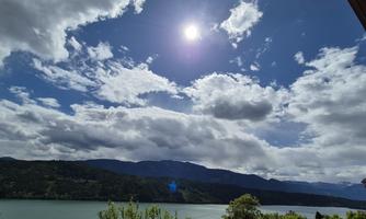 A picturesque view of a lake, surrounded by mountains. The sky is partly cloudy with radiant sunshine.