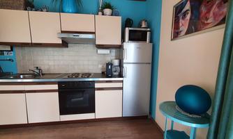 A modern kitchen with blue walls and light furniture. A refrigerator is on the countertop and a cooktop is visible.