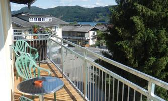 A balcony with green chairs and a small table. In the background, there are houses and trees as well as a clear blue sky.