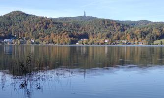 A quiet lake surrounded by colorful trees in autumn. In the background, a mountain rises with a lookout tower.
