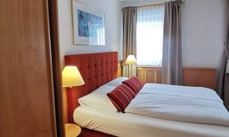 A cozy bedroom with a red bed and white bedding. The room has large windows and a wooden floor.
