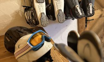 Ski equipment, consisting of ski boots and a helmet with ski goggles, is neatly stored. The ski boots hang on a wall holder, and the helmet is on a wooden shelf.