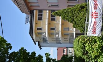 A modern residential building with balconies and a landscaped front garden. In the background, there are more houses and a clear blue sky.