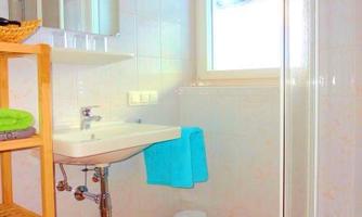 A modern bathroom with a shower and a sink. There is a bright window front and a shelf with towels.
