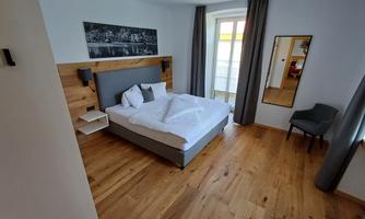 A modern bedroom with a large bed, wooden floor and soft colors. A large mirror and a chair complement the cozy atmosphere.