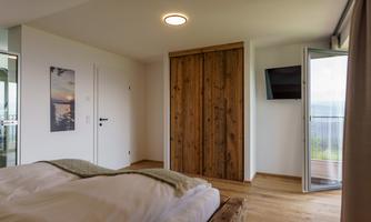 A modern bedroom with a large bed and wooden floorboards. A wooden door and a television on the wall complete the cozy atmosphere.