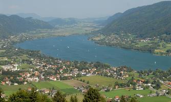 A picturesque landscape with a calm lake and gentle hills. In the foreground, green fields and small houses can be seen.