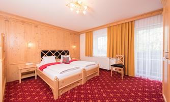 A cozy bedroom with wooden paneling and a large double bed. There are curtains in warm colors and a chair against the wall.