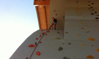 A climber stands at a climbing wall under a wooden roof. The wall is equipped with colored holds in various shapes.