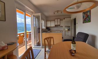 A bright kitchen with a dining table and chairs. In the background, there are windows with a view of the landscape.