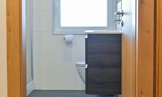 A modern bathroom with a window, a toilet, and a sink. The walls are light-tiled, and the floor is made of dark material.