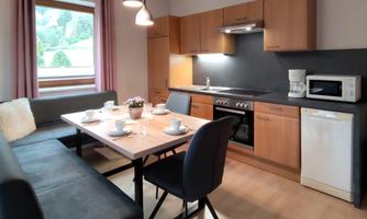 A modern kitchen with wooden cabinets and a dining area. The table is set and there are large windows overlooking greenery.