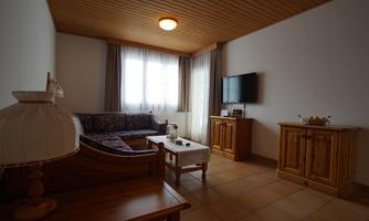 A cozy living room with a sofa, a television, and wooden furniture. Large windows let in a lot of daylight.