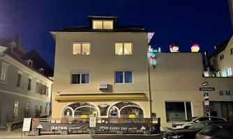 A three-story building in the darkness with glowing windows. In front of the building is a restaurant with a terrace.