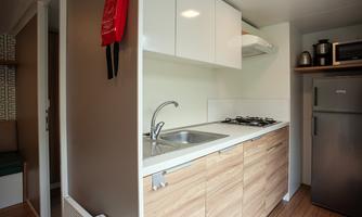 A modern kitchenette with a sink, a stove, and a refrigerator. The floor is made of wood and the walls are kept light.