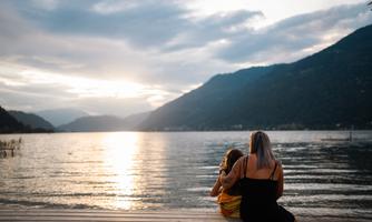 A woman is sitting with a child by the shore of a lake. In the background, mountains and a sunset are visible.