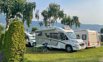 A motorhome is located at a campsite with green trees and a cozy seating area. In the background, more vehicles and mountains can be seen.
