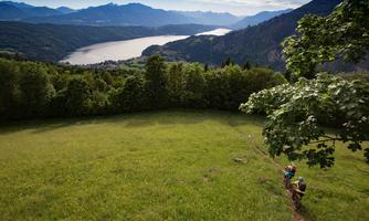 A picturesque landscape with a river and mountains in the background. Hikers explore the green hill under a clear sky.