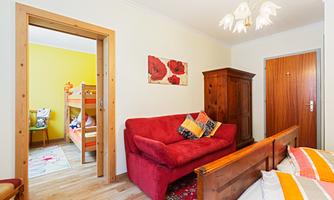 A cozy room with a red sofa and a wooden wardrobe. In the background, there is a children's room with a bunk bed and yellow wall color.