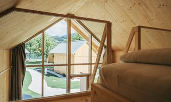 A cozy attic space with a wooden ceiling and a large window overlooking a green landscape. In the foreground, there is a bed with soft pillows visible.
