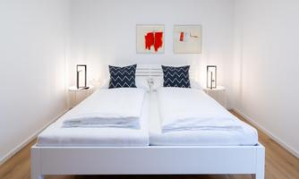 A modern bedroom with a large white bed and blue pillows. Two simple artworks hang on the walls, and the lighting is soft.