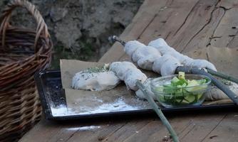 A rustic wooden table with fresh, uncooked bread stuck on sticks. Next to the bread is a bowl of fresh vegetables and spices.