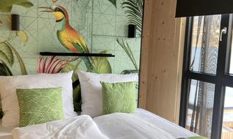 A modern bedroom with a green accent on the wall and decorative pillows. It features a cozy bed and a large window for natural lighting.