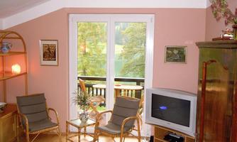 A cozy living room with rattan-like chairs and a television. Large windows offer a view of the green landscape outside.