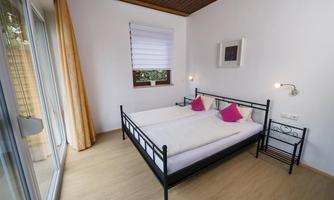 A modern bedroom with a large double bed and pink pillows. Large windows provide plenty of daylight and a view outside.