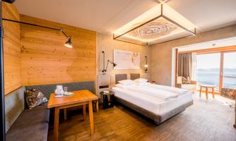 A modern hotel room with a double bed, a table, and a cozy seating area. Large windows offer a view of the surrounding landscape.