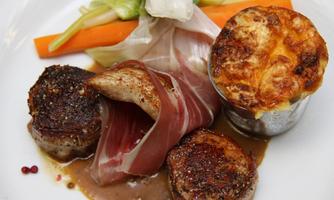 A carefully arranged dish with fried meat, wrapped in ham, and a side of vegetables. There is also a small portion of casserole in a metal dish.