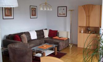 A cozy living room with a large brown sofa and a glass table. Pictures hang on the walls, and a bookshelf stands in the corner.