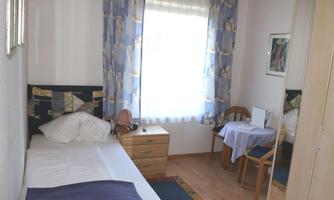 A simple single room with a bed, a table, and two chairs. Bright curtains let in plenty of daylight.