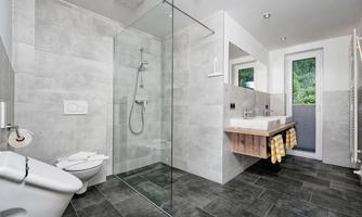 A modern bathroom with a glass shower and a large window front. The floor is tiled with dark tiles and there is a wooden shelf with towels.