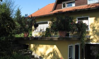 A well-maintained house with a colorful balcony full of plants. The garden around the house is green and inviting.