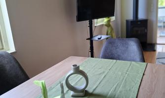 A modern living room with a wooden table and green tablecloth. In the background, there is a television and some decorative elements on the wall.